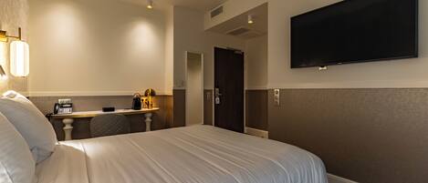 A hotel room with a large bed, a desk with a lamp, a telephone, and a TV mounted on the wall.