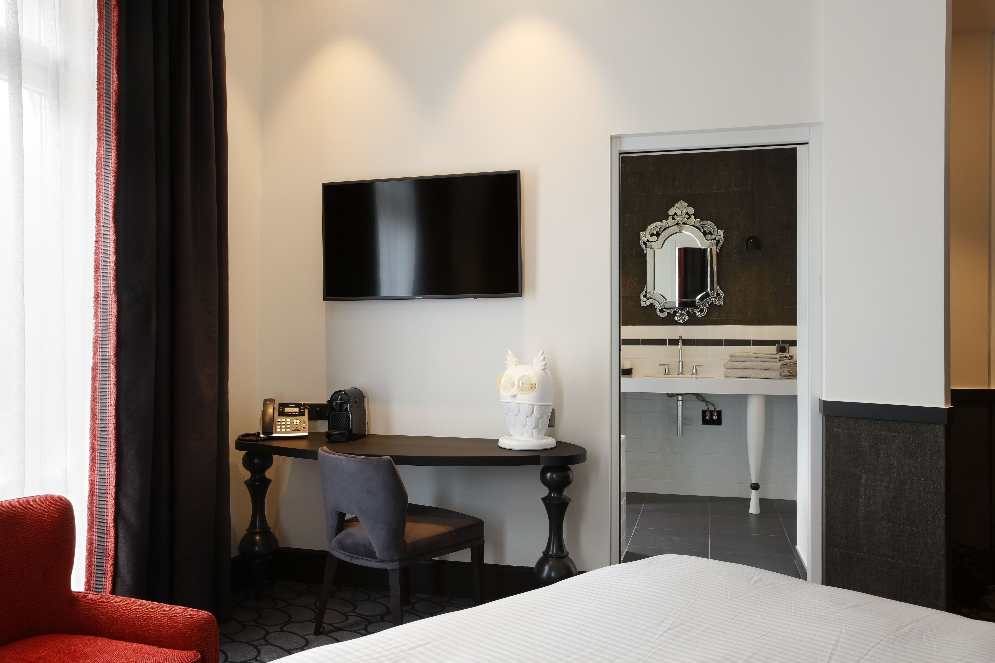 A hotel room with a bed, a desk with a chair, a television, a mirror, and a bathroom visible through an open door.