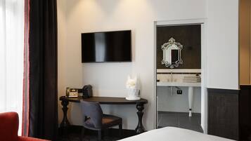 Habitación de hotel con cama, escritorio con silla, televisión, espejo y baño visible a través de una puerta abierta.