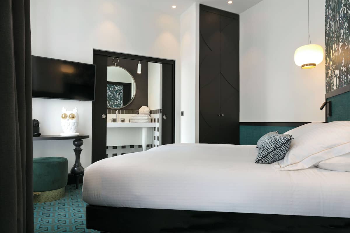 Una habitación de hotel moderna con una cama grande, un televisor montado en la pared, un espejo redondo y una mesita de noche con una lámpara y objetos decorativos.