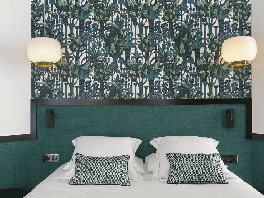 A hotel room with a patterned wallpaper, a bed with white bedding and patterned pillows, two wall-mounted lamps, and a green upholstered headboard.