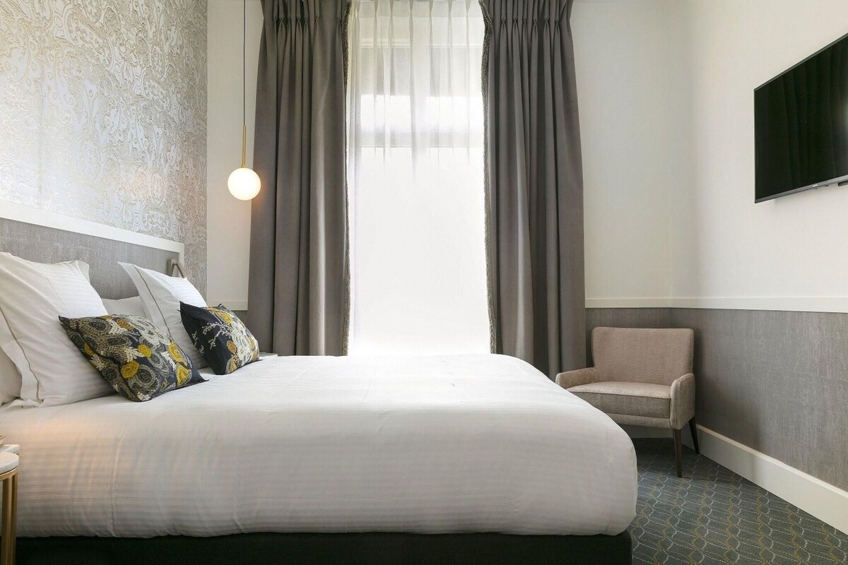 A hotel room with a large bed, a chair, a TV mounted on the wall, and a window with curtains.