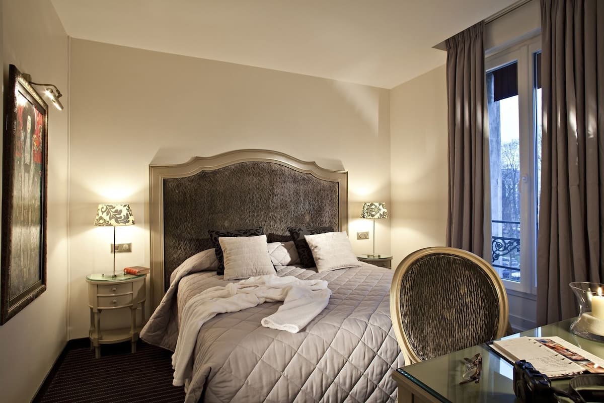 Una habitación de hotel con una cama grande, dos lámparas de noche, una silla, una mesa con una vela y una ventana con cortinas.