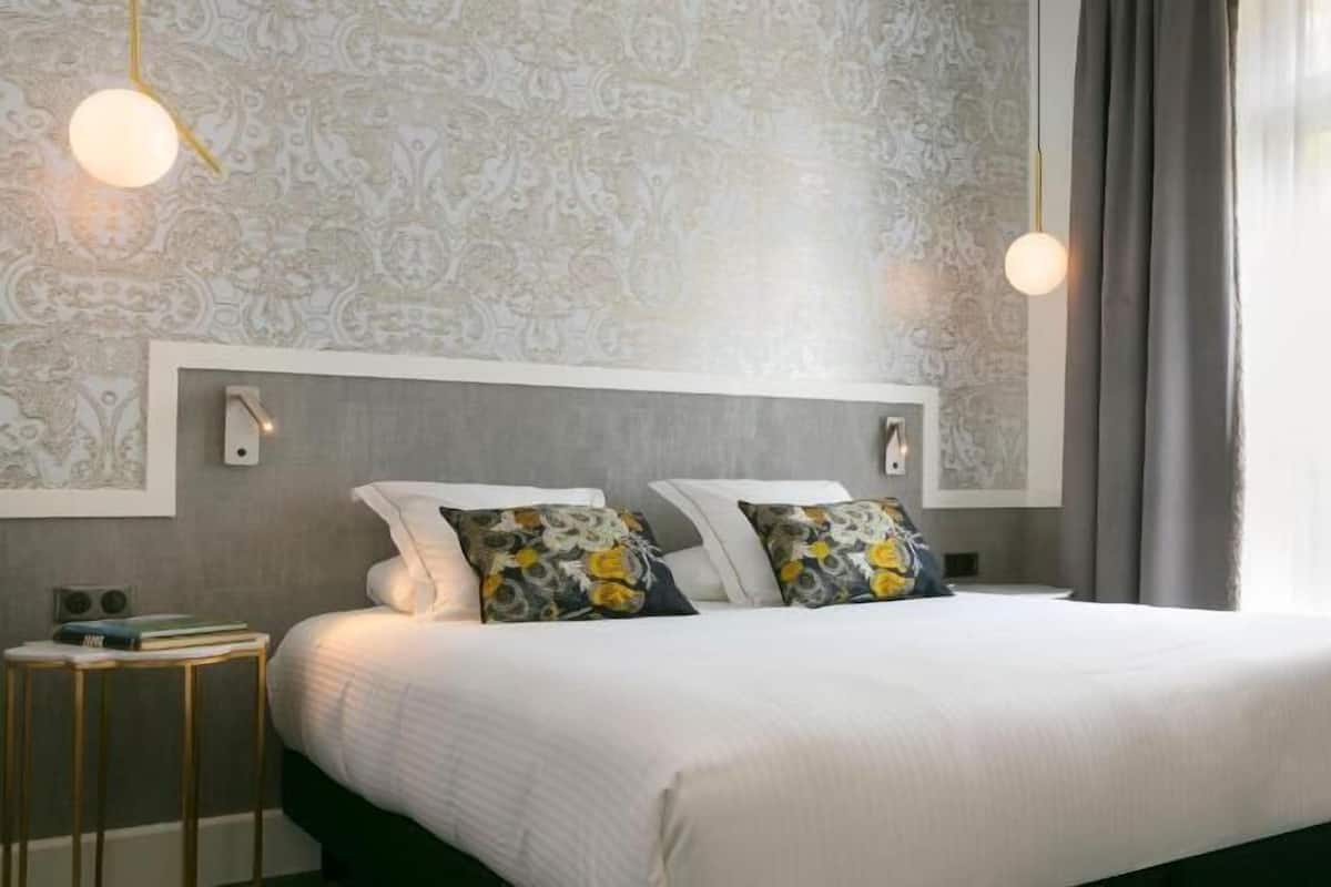 Habitación de hotel con una cama grande, dos lámparas de pared, una mesita de noche pequeña y una ventana con cortinas.
