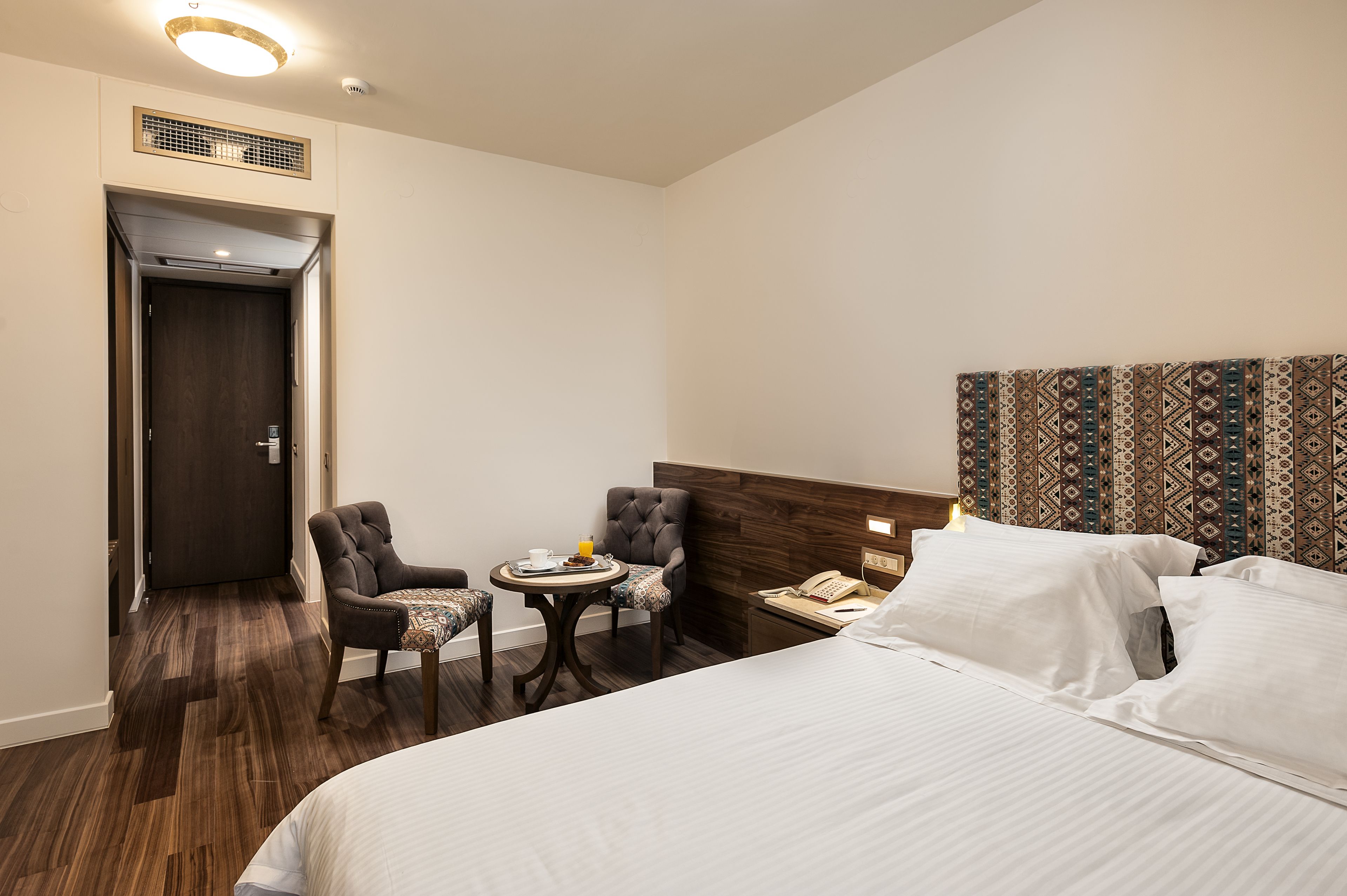 A hotel room with a bed, a small table with a chair, a telephone, and a patterned headboard.
