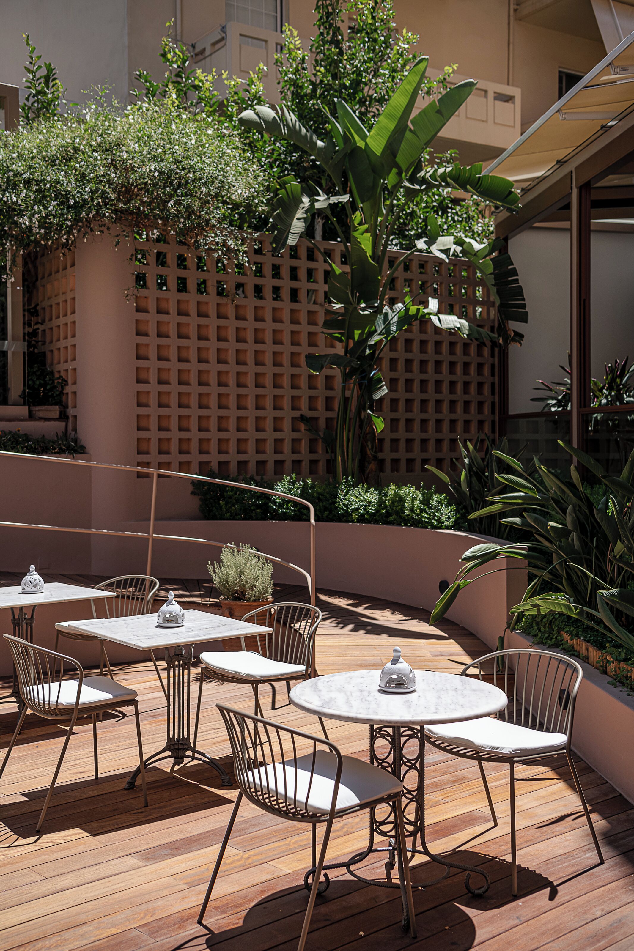Un espacio de patio al aire libre con mesas redondas y sillas de metal, rodeado de abundante vegetación y un cerramiento de celosía.