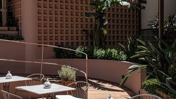Un espacio de patio al aire libre con mesas redondas y sillas de metal, rodeado de abundante vegetación y un cerramiento de celosía.