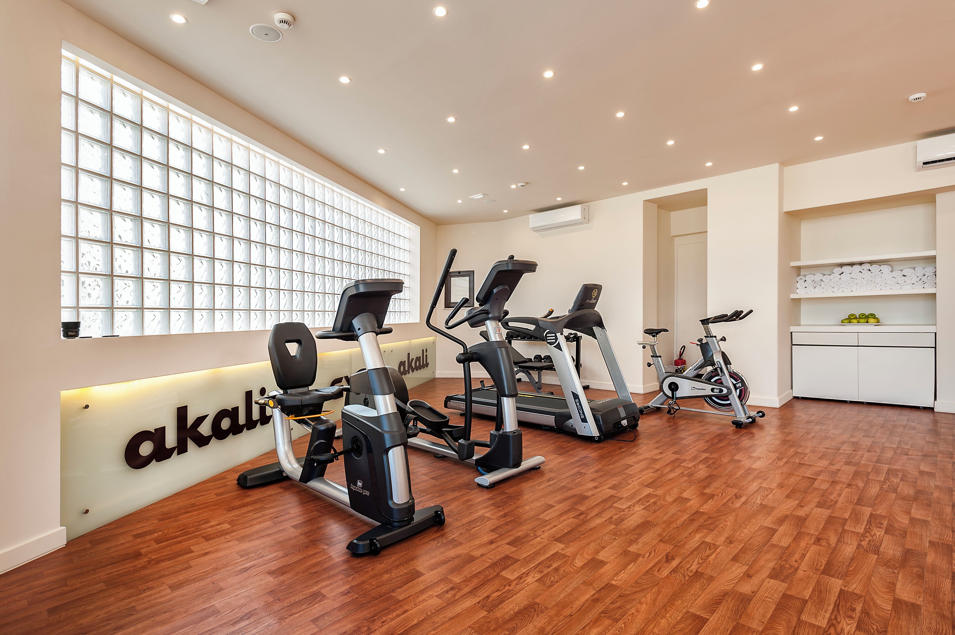 Een sportschool met meerdere fitnessapparaten, een groot raam met zonwering en een witte muur met een plank.