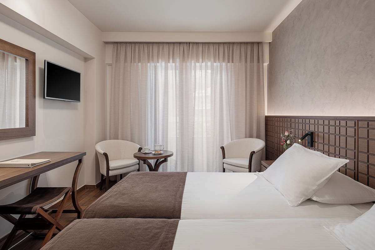Ein Hotelzimmer mit einem Bett, zwei Stühlen, einem kleinen Tisch mit Teekanne und Tassen, einem an der Wand befestigten Fernseher und einem Fenster mit Vorhängen.