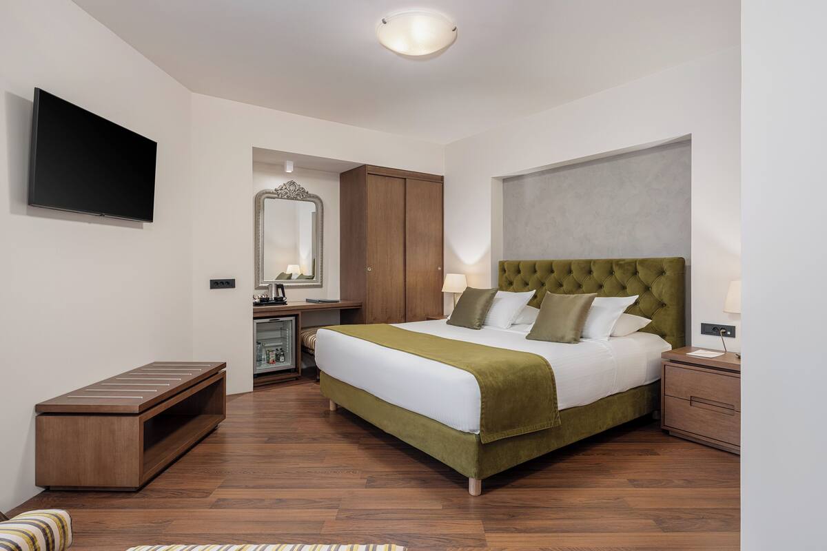 Ein modernes Hotelzimmer mit einem großen Bett, einem Flachbildfernseher, einer hölzernen Sitzbank, einem Spiegel und einem Kleiderschrank.