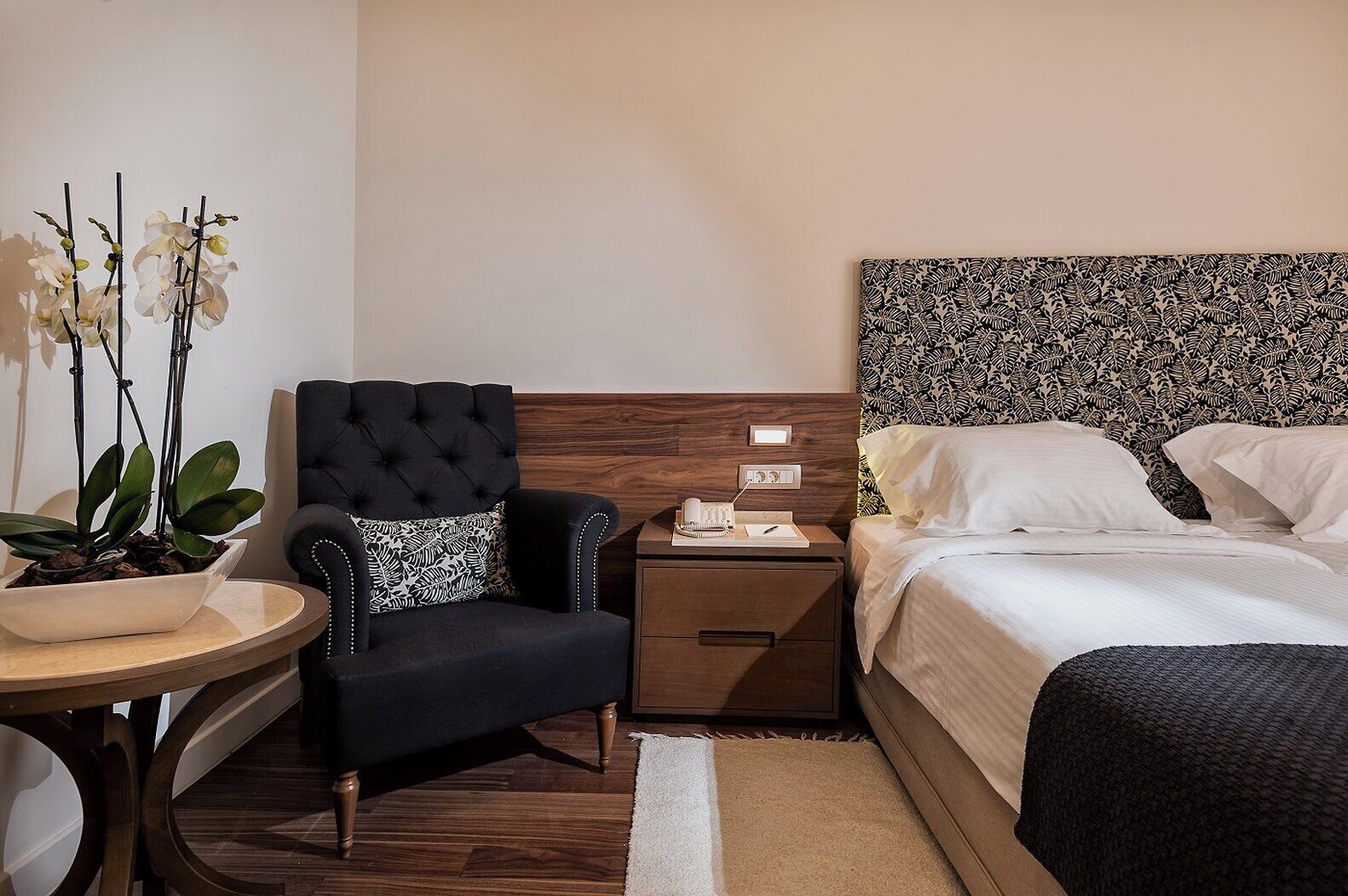 A hotel room with a bed, a nightstand, a chair, a small table with a plant, and a headboard with a leaf pattern.