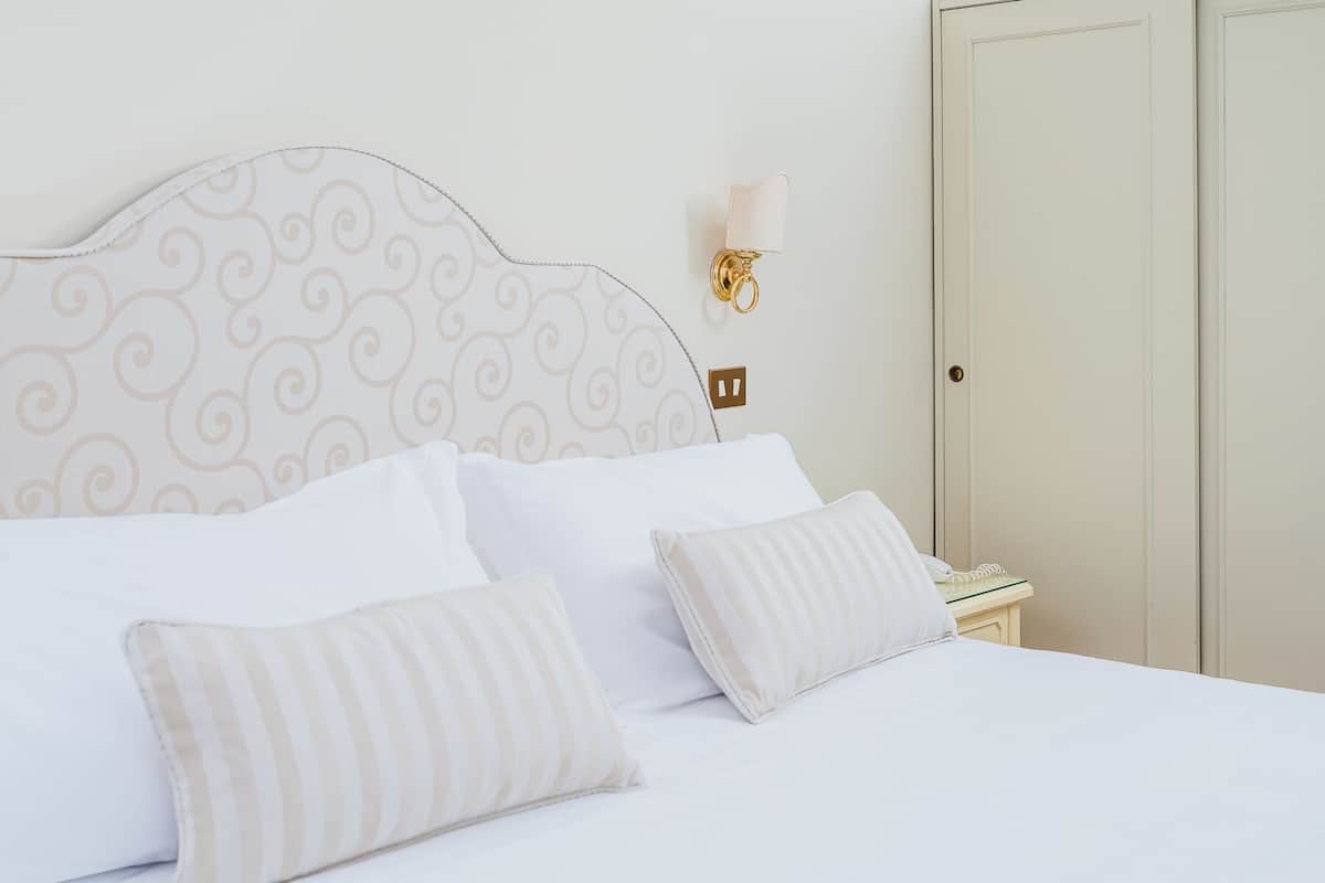 Un lit bien fait, avec du linge de lit blanc et des oreillers à rayures, un luminaire fixé au mur et une porte fermée, dans une chambre d’hôtel.