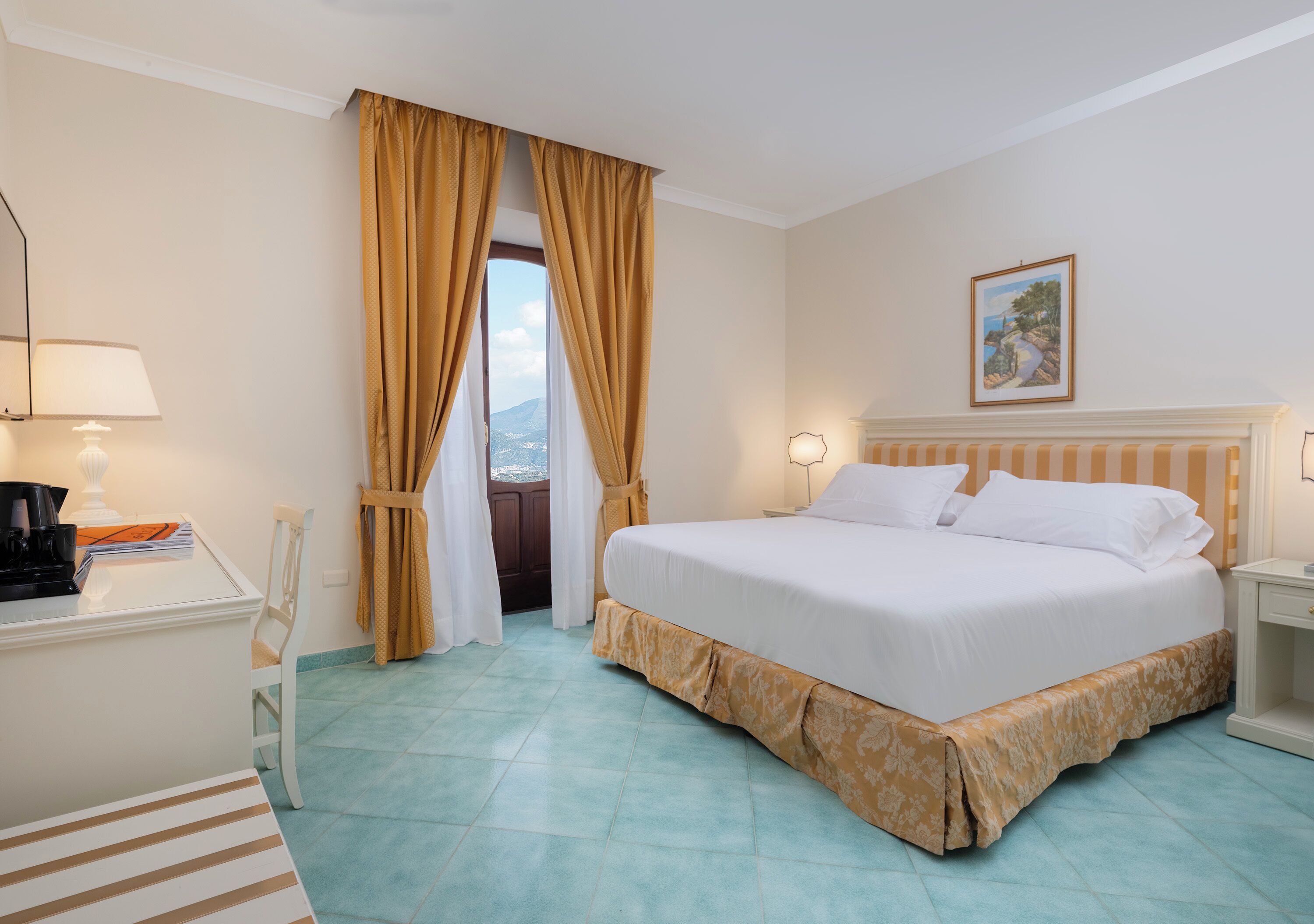 Una habitación de hotel con cama, escritorio con lámpara, silla, ventana con cortinas y un cuadro en la pared.