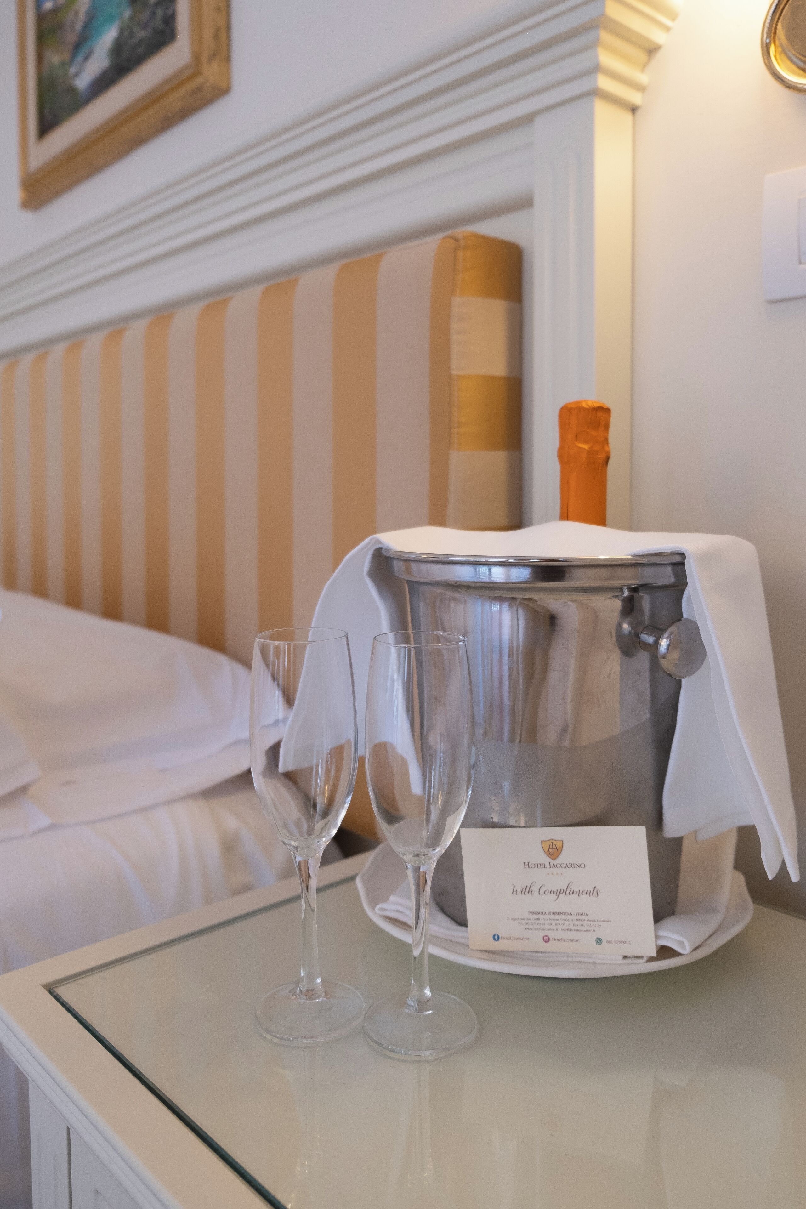 Una habitación de hotel con una cama, una mesita de noche con cubero y copas de champán, y un cuadro enmarcado en la pared.