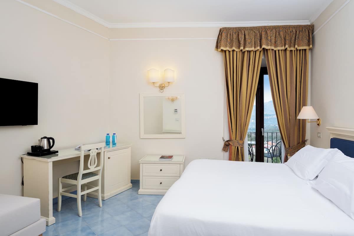 Une chambre d’hôtel comprenant un lit, un bureau avec une chaise, une commode, un miroir, une télévision, une fenêtre avec des rideaux et un balcon avec vue.