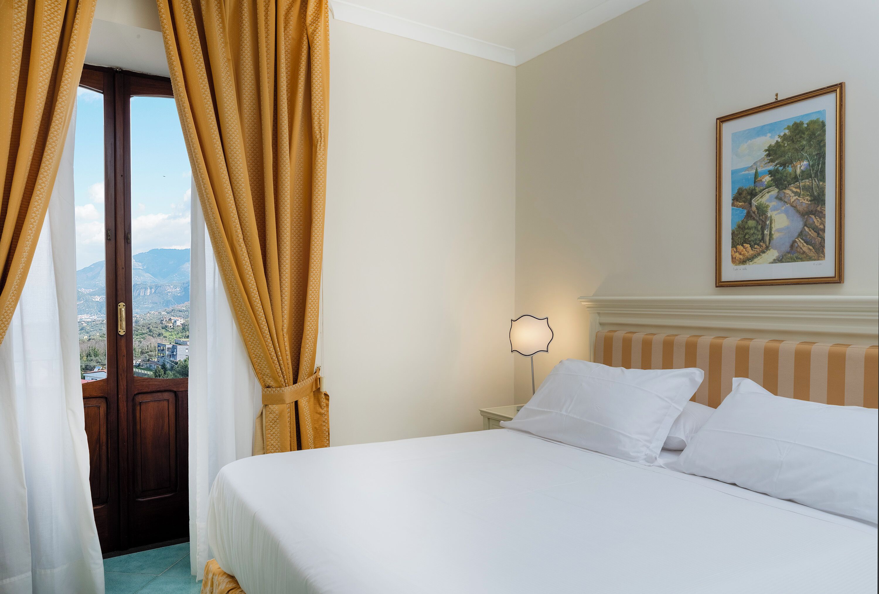 Une chambre d’hôtel avec un lit, une table de chevet, une lampe, un tableau et une vue sur l’extérieur, visible à travers une porte.