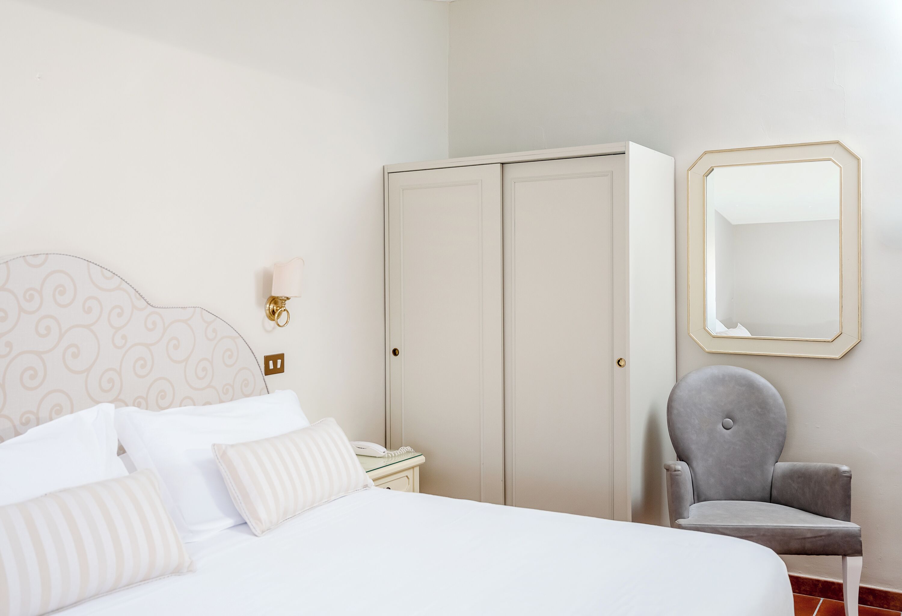 A bedroom with a bed, a wardrobe, a chair, a bedside table, and a wall-mounted mirror.