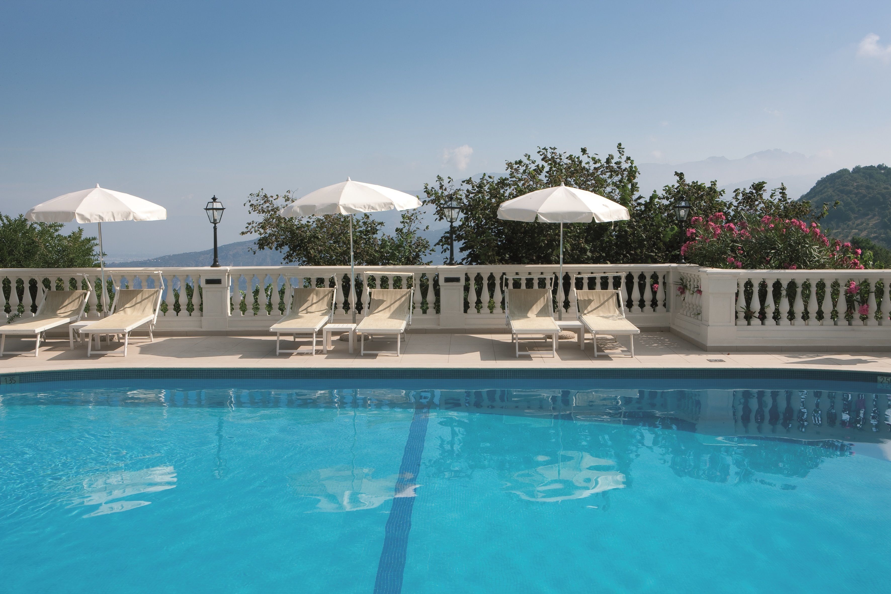A pool with white lounge chairs and umbrellas, surrounded by a white balustrade and greenery.