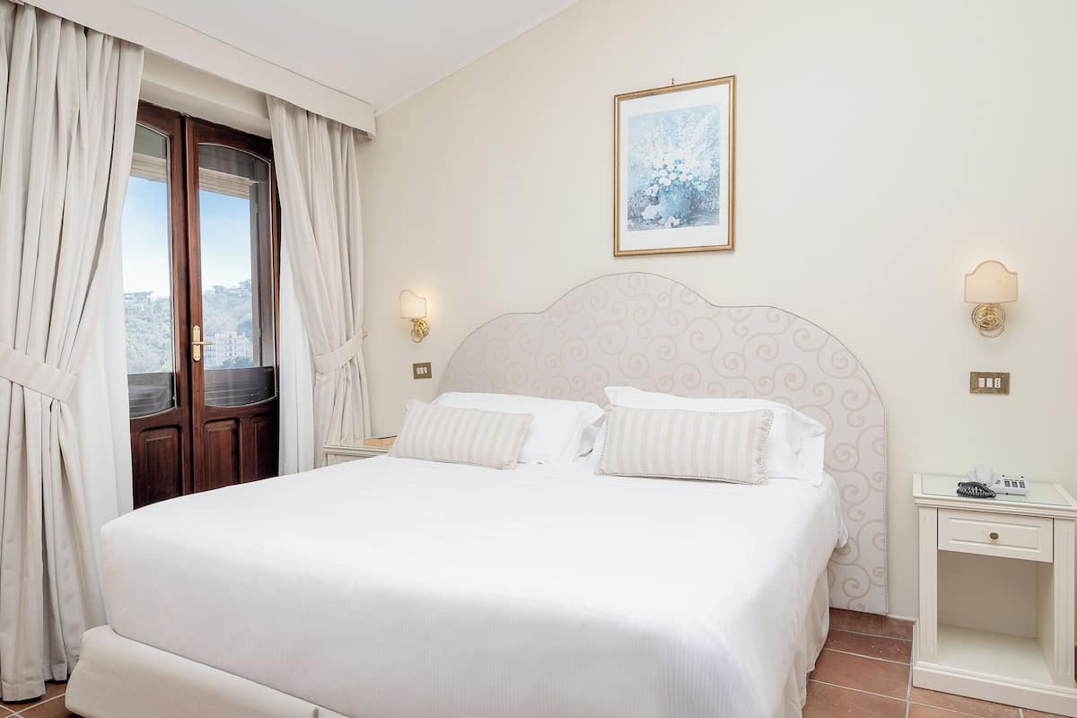 Une chambre d’hôtel avec un lit, des rideaux, une vue par la fenêtre, une lampe fixée au mur et un tableau encadré.
