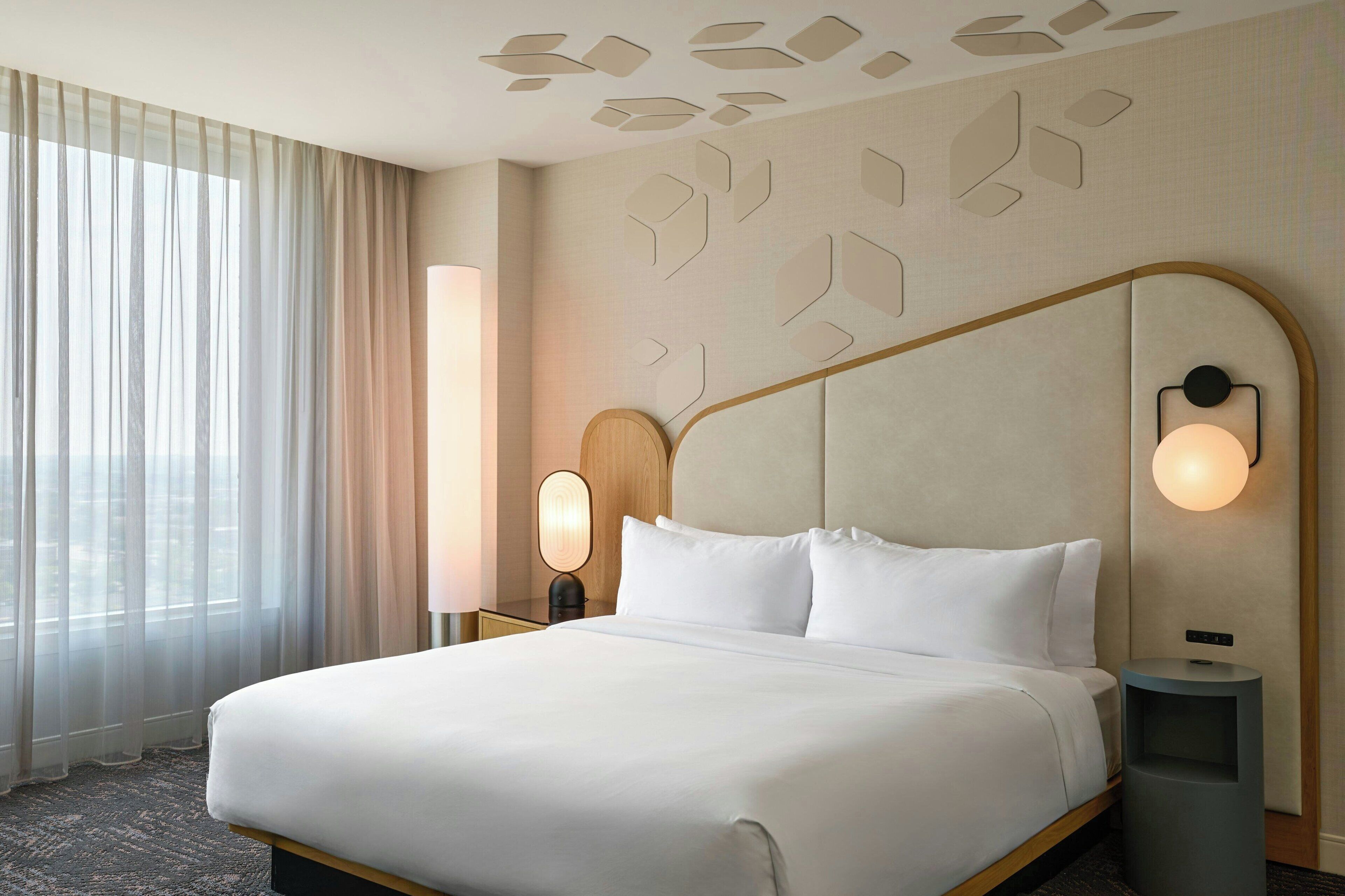 Habitación de hotel con una cama grande, una mesita de noche con lámpara, luz de pared y ventana con cortinas translúcidas.
