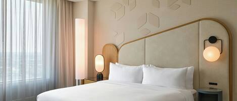 Habitación de hotel con una cama grande, una mesita de noche con lámpara, luz de pared y ventana con cortinas translúcidas.