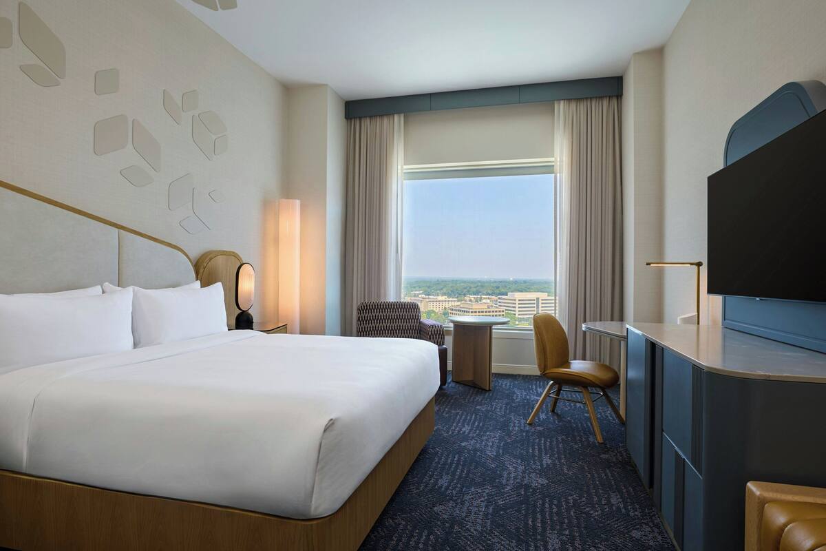 A hotel room with a large bed, a desk, a chair, a TV, and a view of the city.