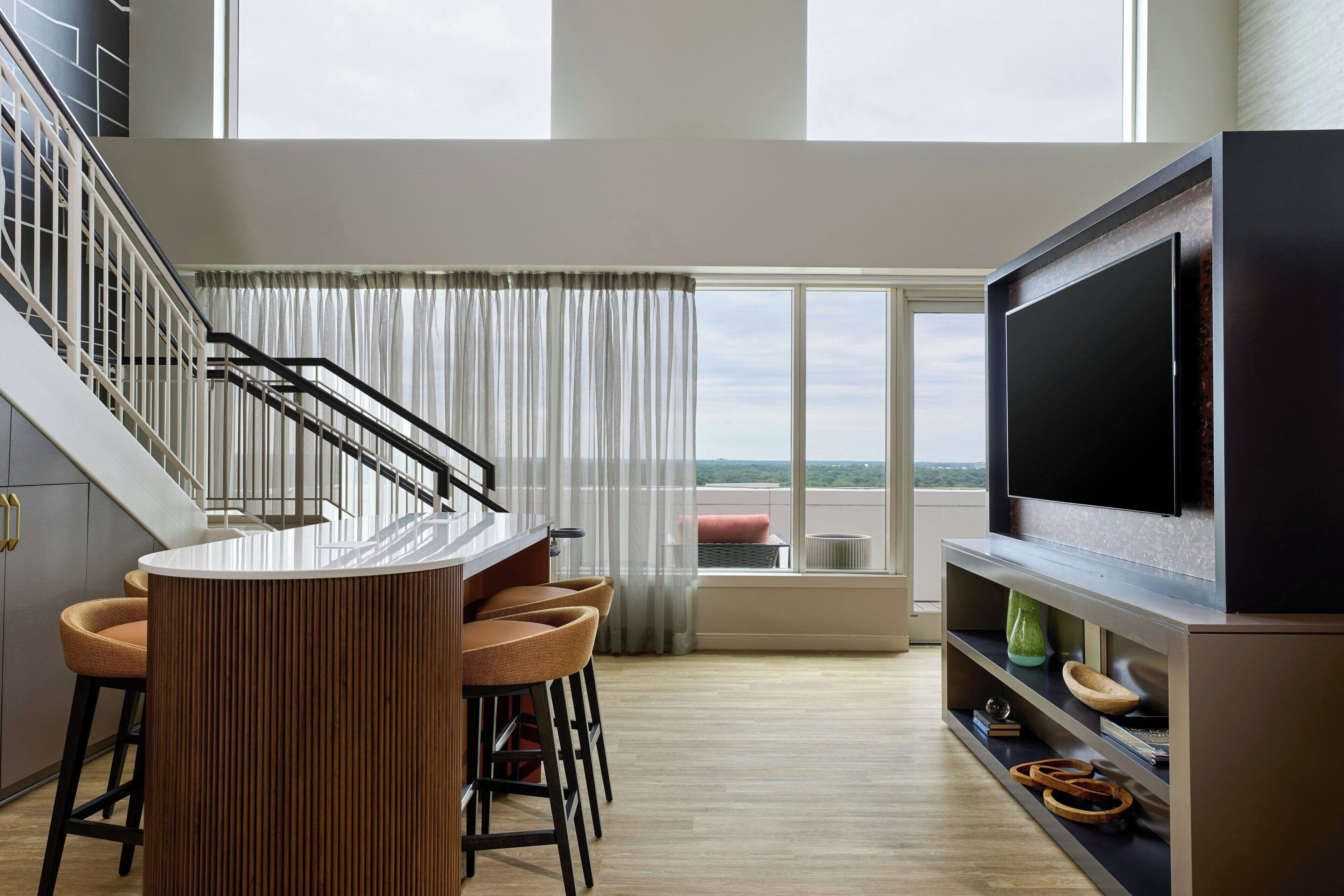 A modern kitchen with a central island, bar stools, a large flat-screen TV, and a staircase leading to an upper level.