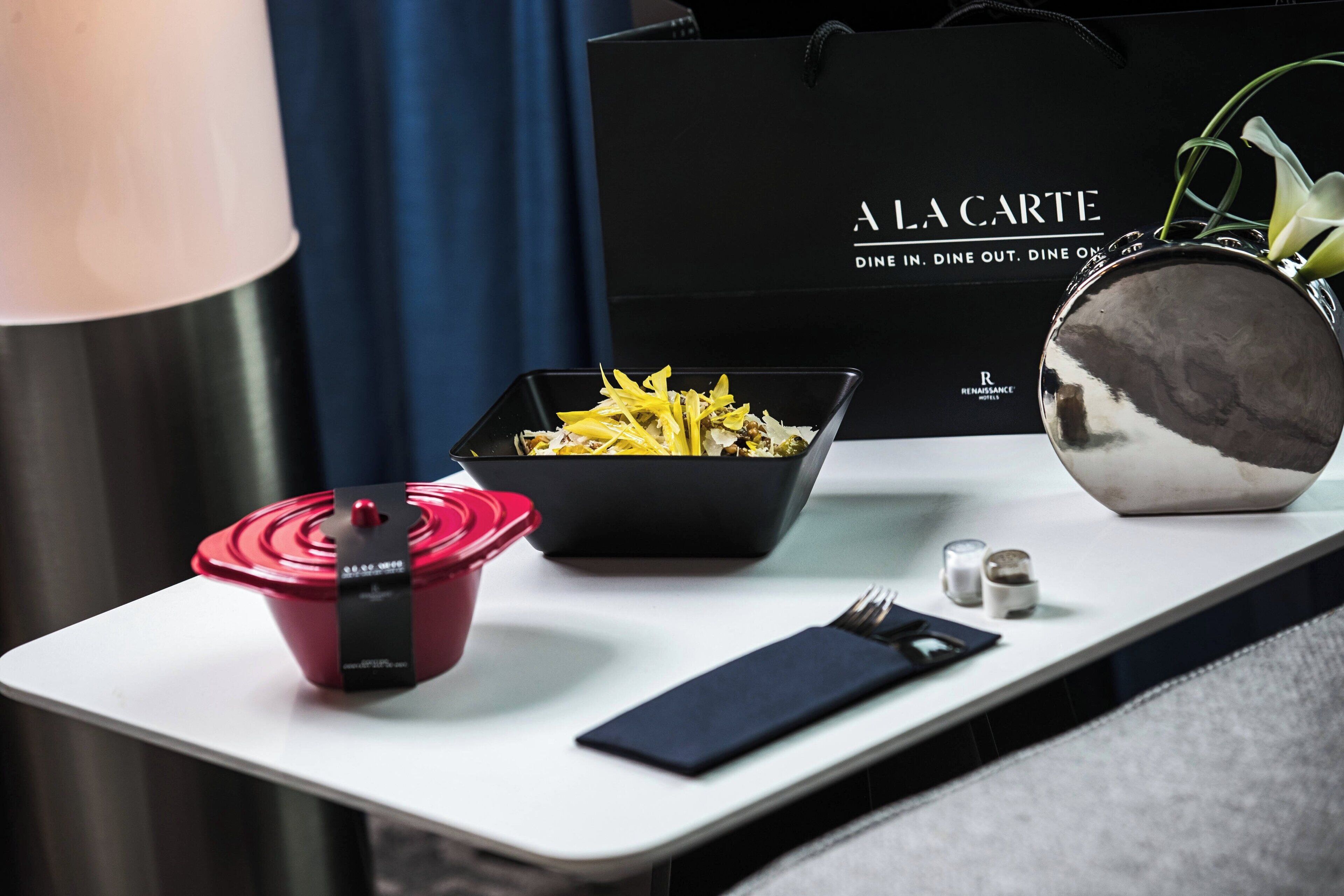 A dining table with a black bowl of food, a red container, a black napkin, and a silver bowl. A black bag with "A LA CARTE" text is in the background.