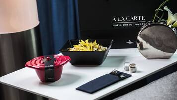 A dining table with a black bowl of food, a red container, a black napkin, and a silver bowl. A black bag with "A LA CARTE" text is in the background.