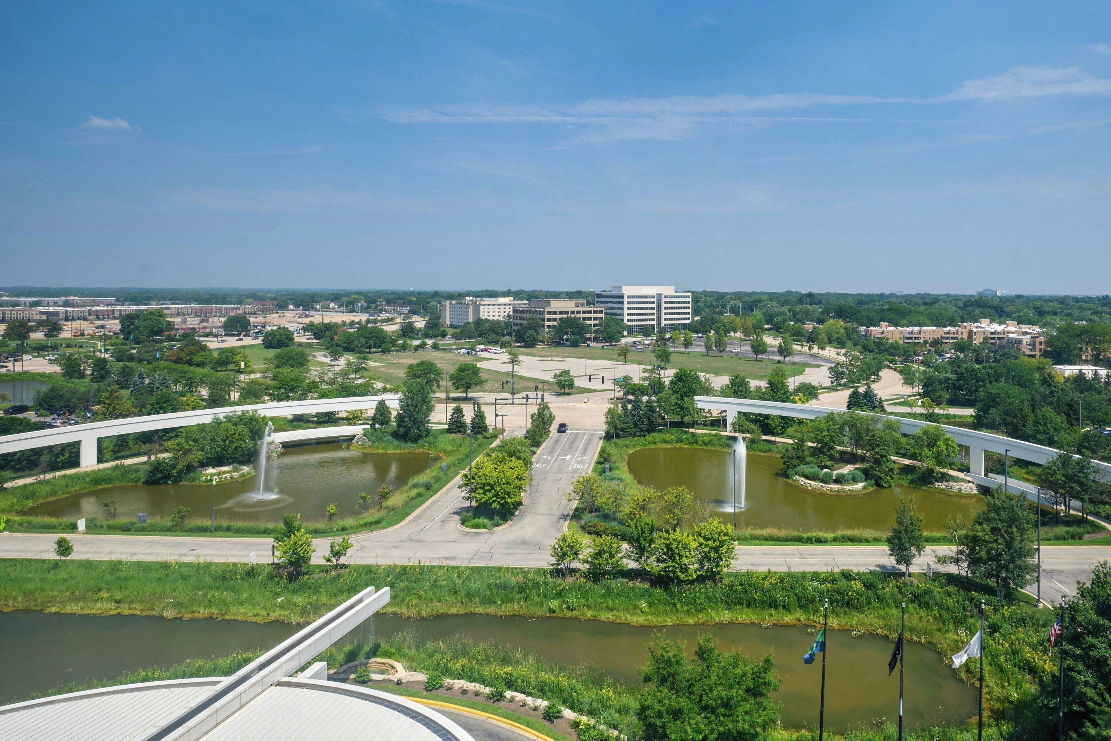 A large, open plaza with a central fountain, surrounded by greenery and a bridge.