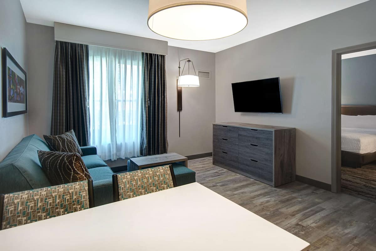 A modern hotel room with a sofa, a TV mounted on the wall, a wooden dresser, and a large window with curtains.