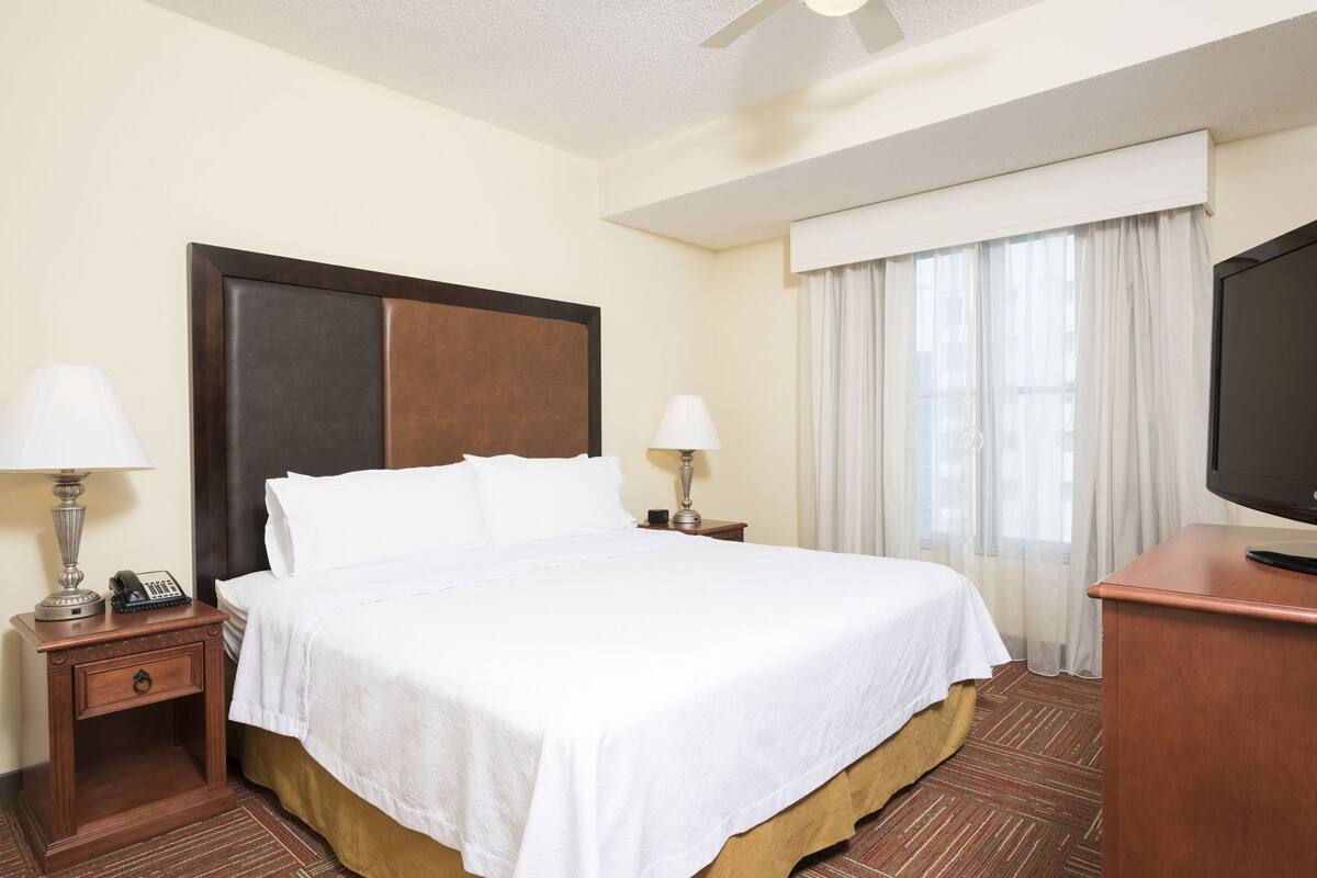 A hotel room with a bed, two bedside tables, a lamp, a telephone, a television, and a window with curtains.