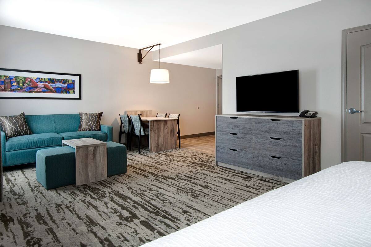 A modern hotel room with a teal sofa, a wooden coffee table, a TV on a dresser, and a painting on the wall.