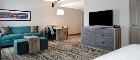 A modern hotel room with a teal sofa, a wooden coffee table, a TV on a dresser, and a painting on the wall.