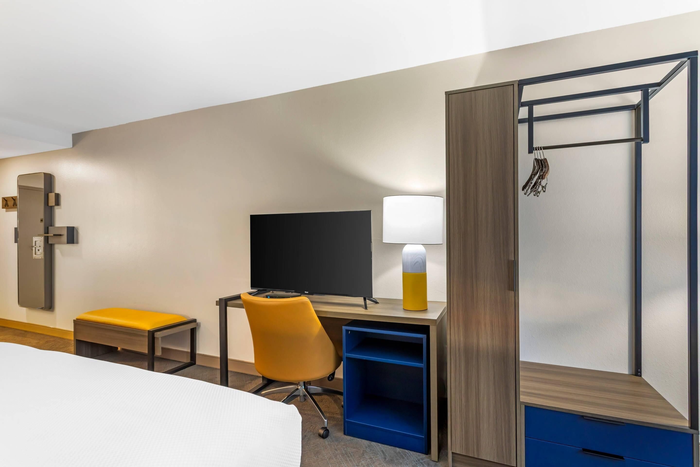 A hotel room with a bed, a desk with a chair, a television, a lamp, a wardrobe, and a coat rack.