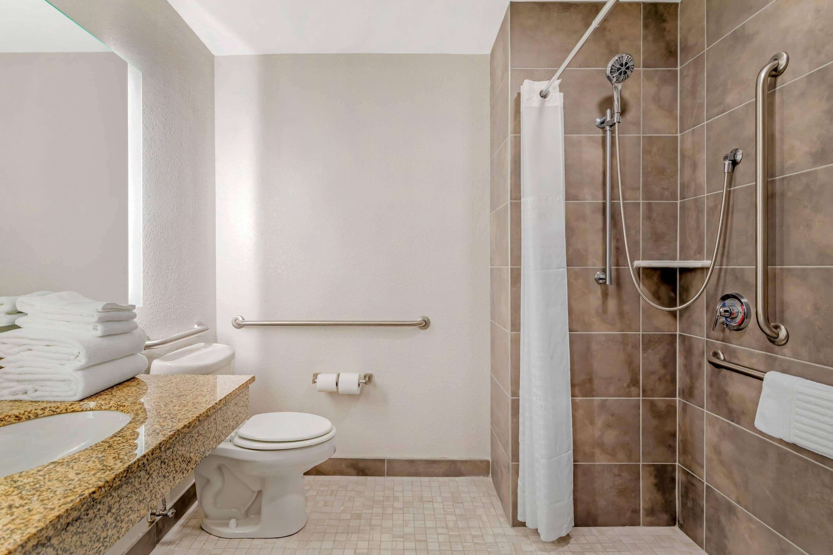 Un baño moderno con encimera de granito, inodoro blanco, ducha con cabezal de ducha manual y barra de apoyo.