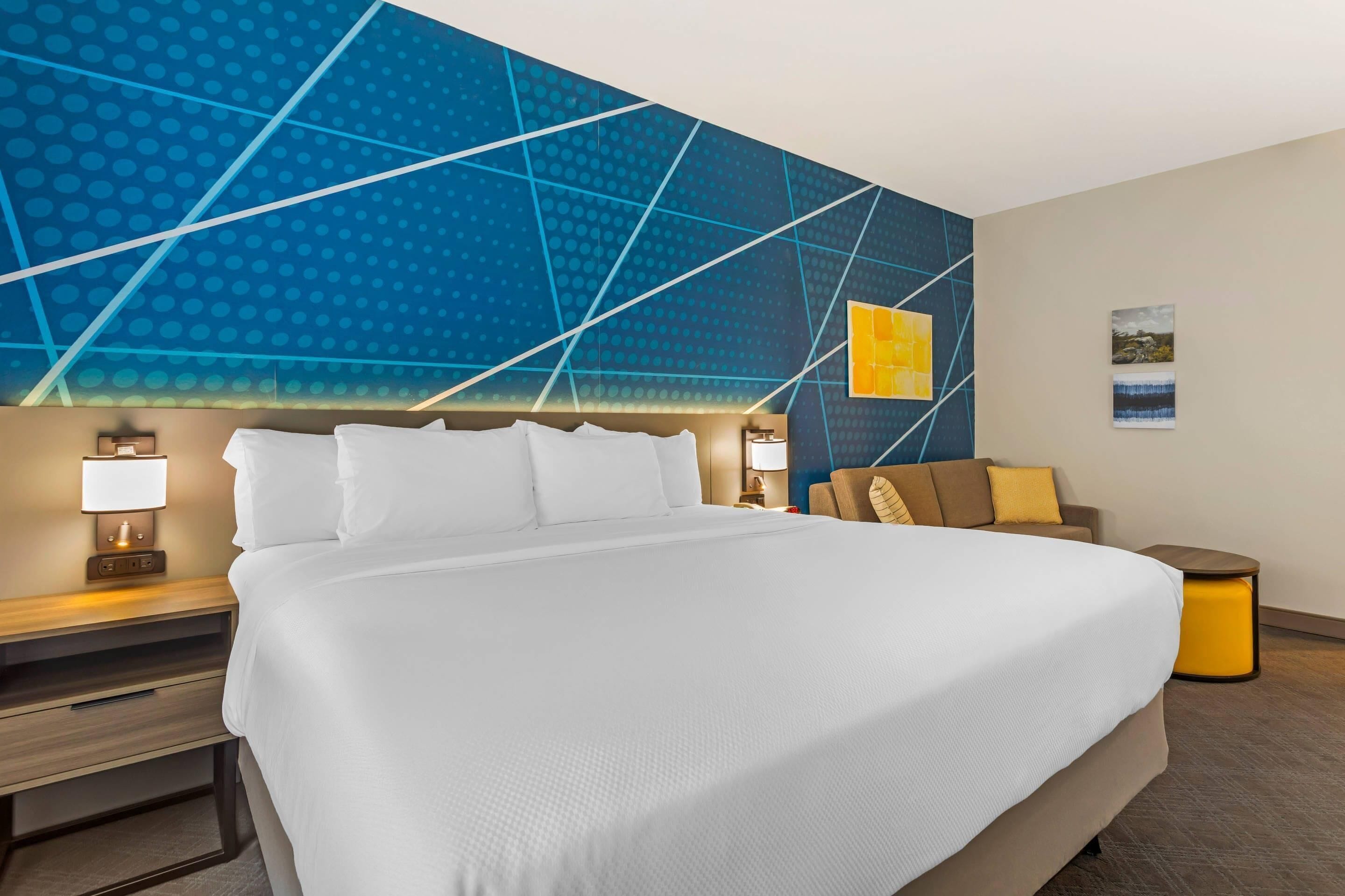 Une chambre d’hôtel avec un grand lit, une table de chevet avec une lampe, un mur orné d’un motif géométrique et une petite table avec une lampe.