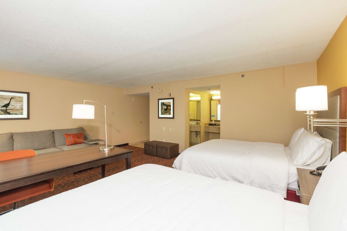 Une chambre d’hôtel avec deux lits, un canapé, un bureau et une salle de bain visible à travers une porte ouverte.