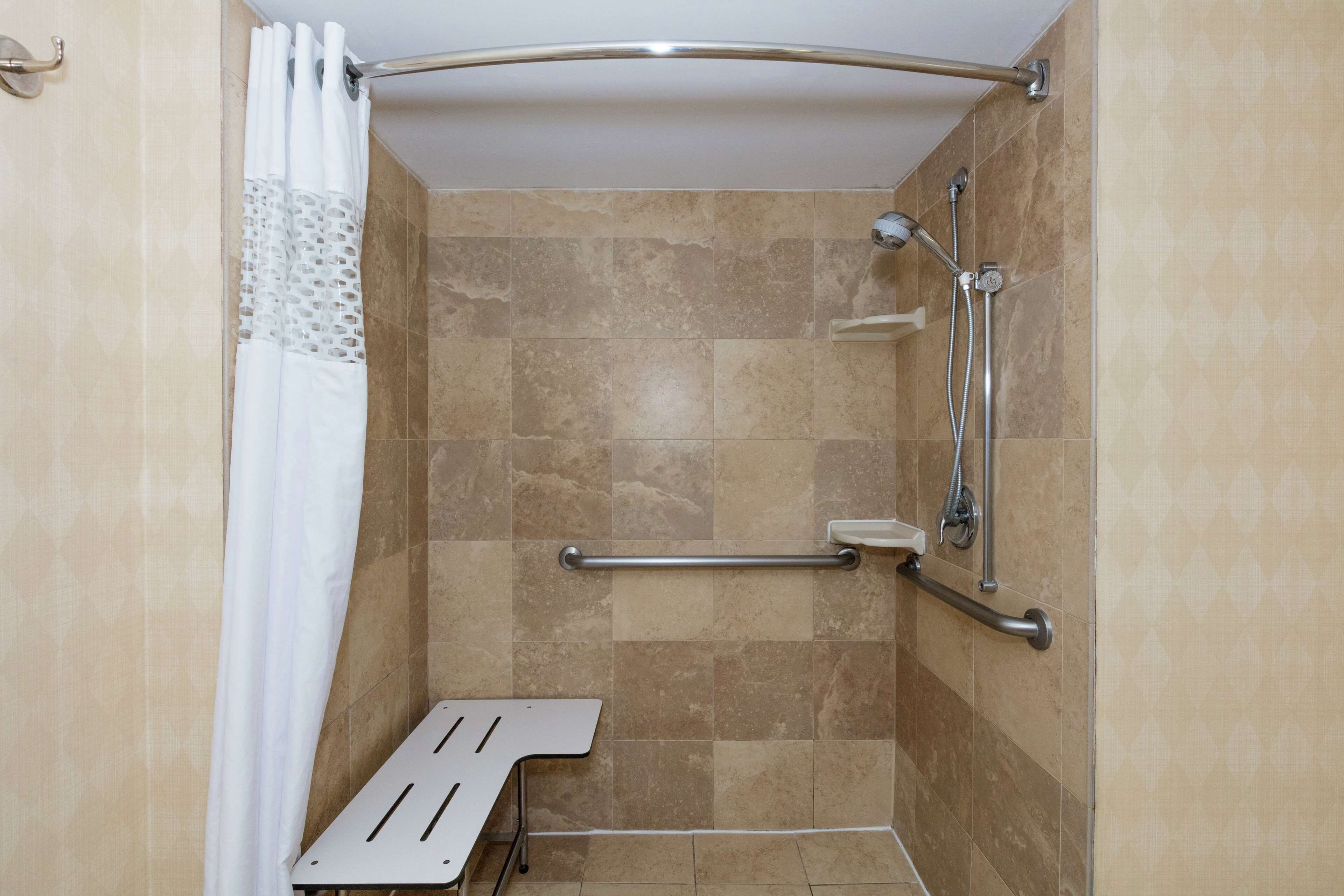 Un espace douche avec des carreaux de couleur beige, une barre d’appui et un siège de douche blanc.