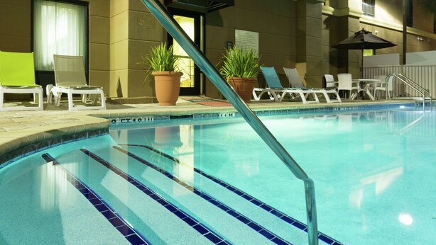 An indoor swimming pool with clear blue water, tiled edges, and a metal ladder. There are lounge chairs and potted plants around the pool area.