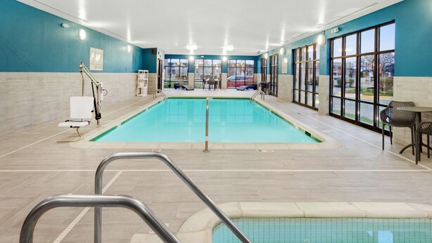 A hotel indoor pool with a clear view of the outside, a lifeguard chair, and a seating area with tables and chairs.