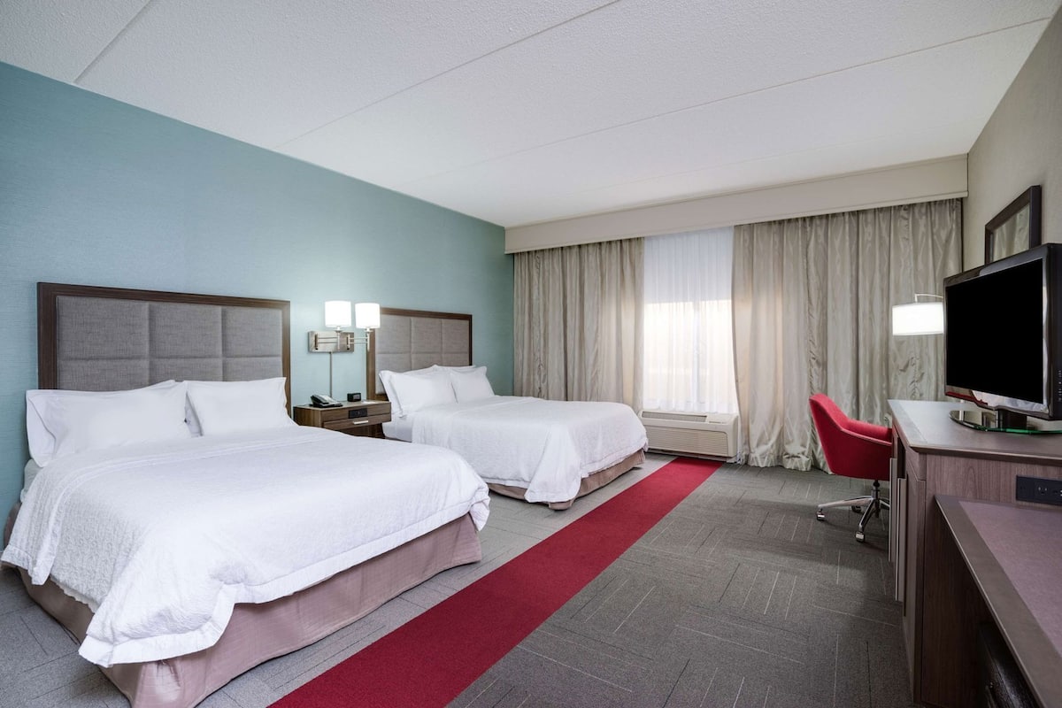 Habitación de hotel con dos camas, un escritorio con televisión, una silla, una lámpara de escritorio y una ventana con cortinas.