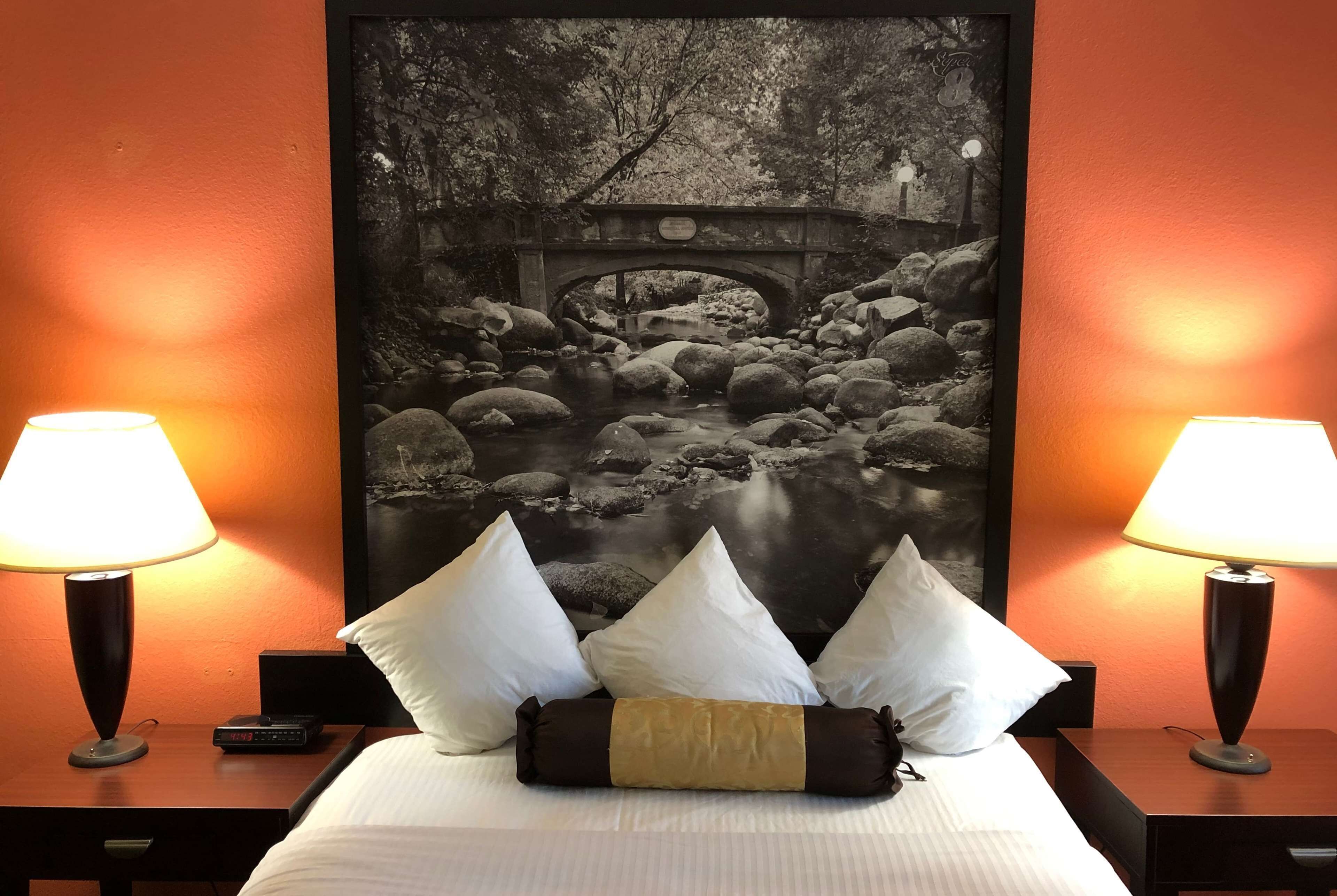 Ein Hotelzimmer mit einem Bett, weißen Kissen, einem Nachttisch mit Lampe sowie einem Schwarz-Weiß-Foto einer Brücke über einem Bach.