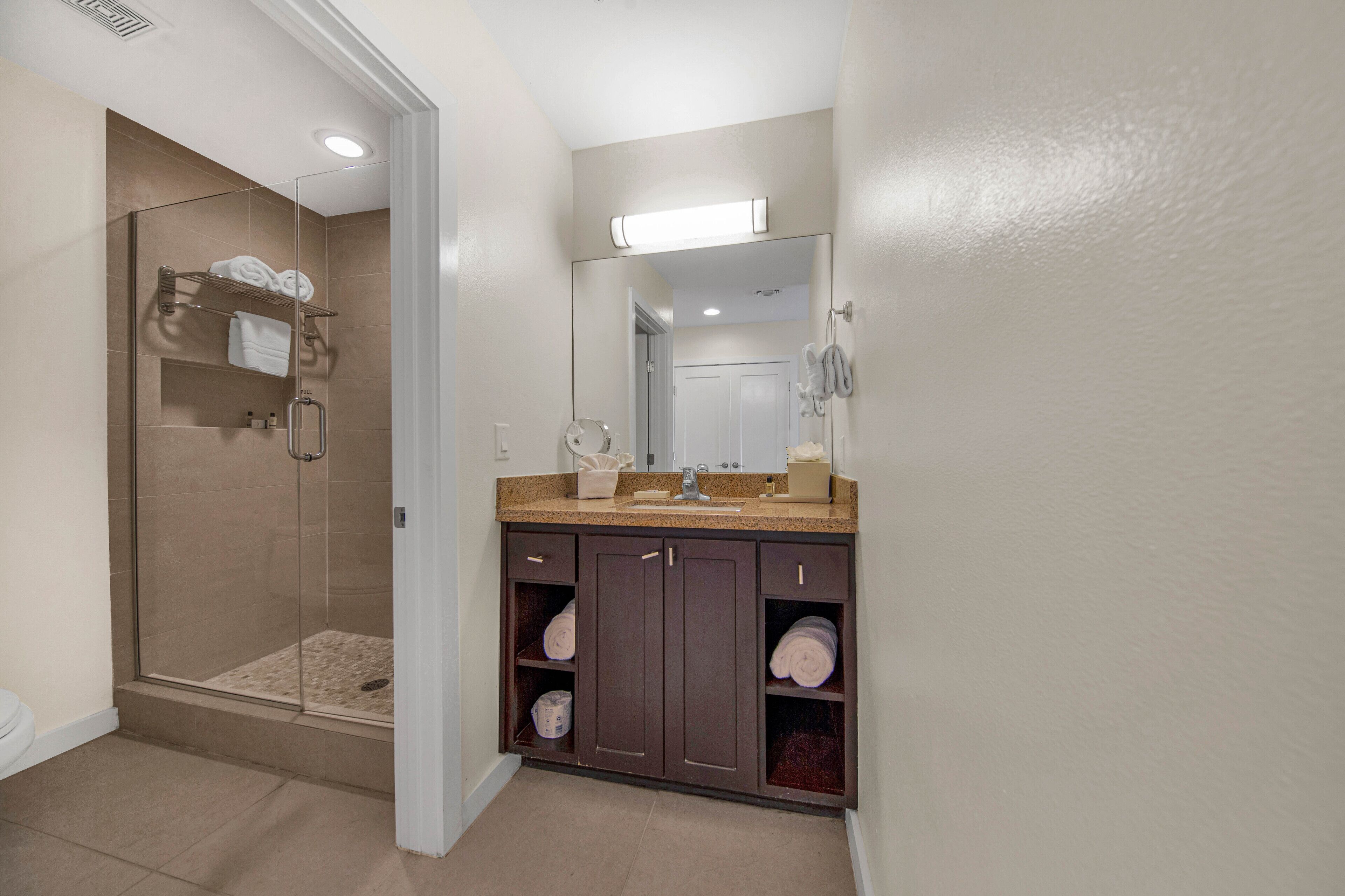 A bathroom with a glass-enclosed shower, a vanity with a mirror, and shelves for towels.