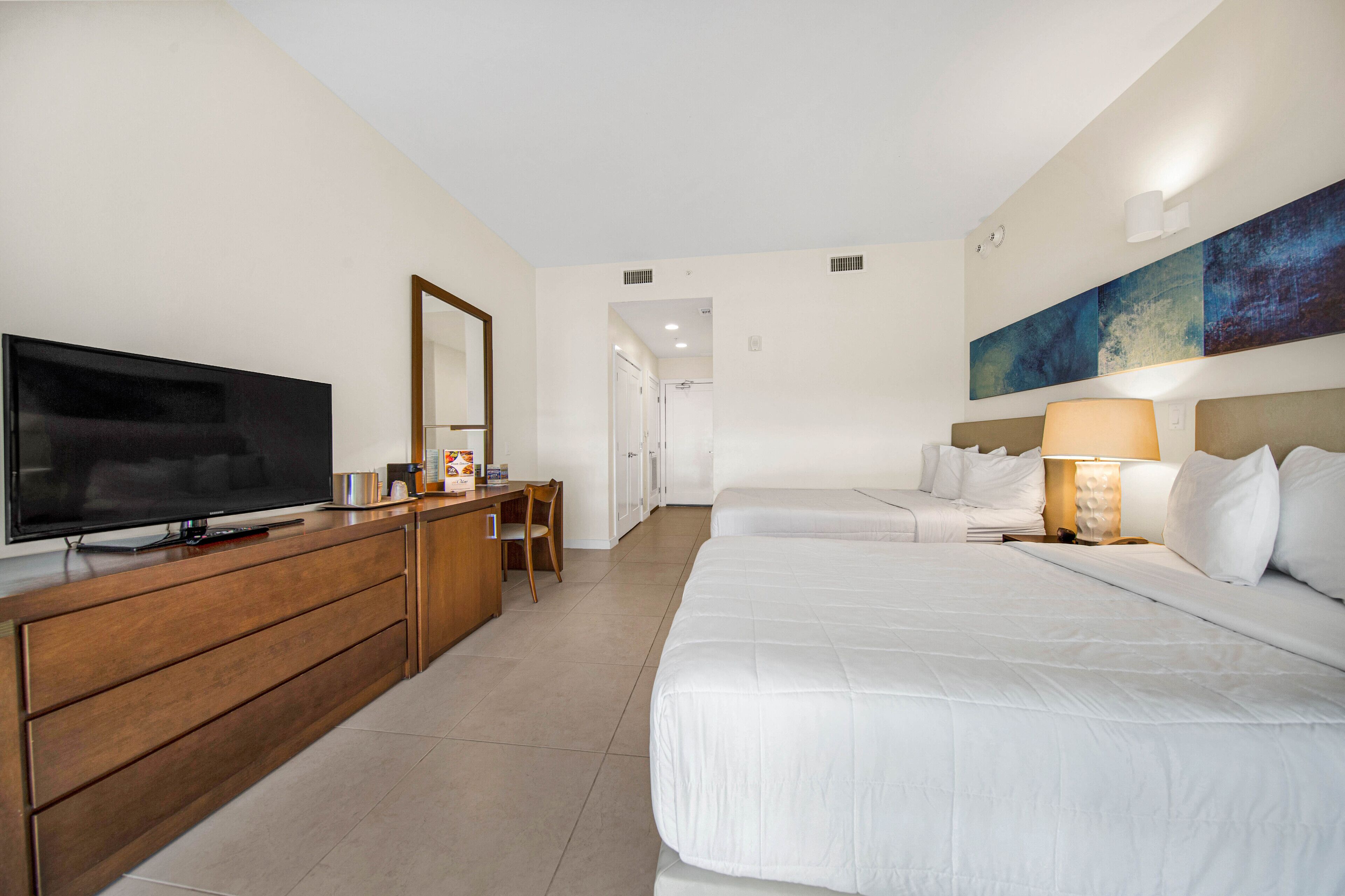 A hotel room with a TV, wooden dresser, two beds, a desk, and a painting on the wall.