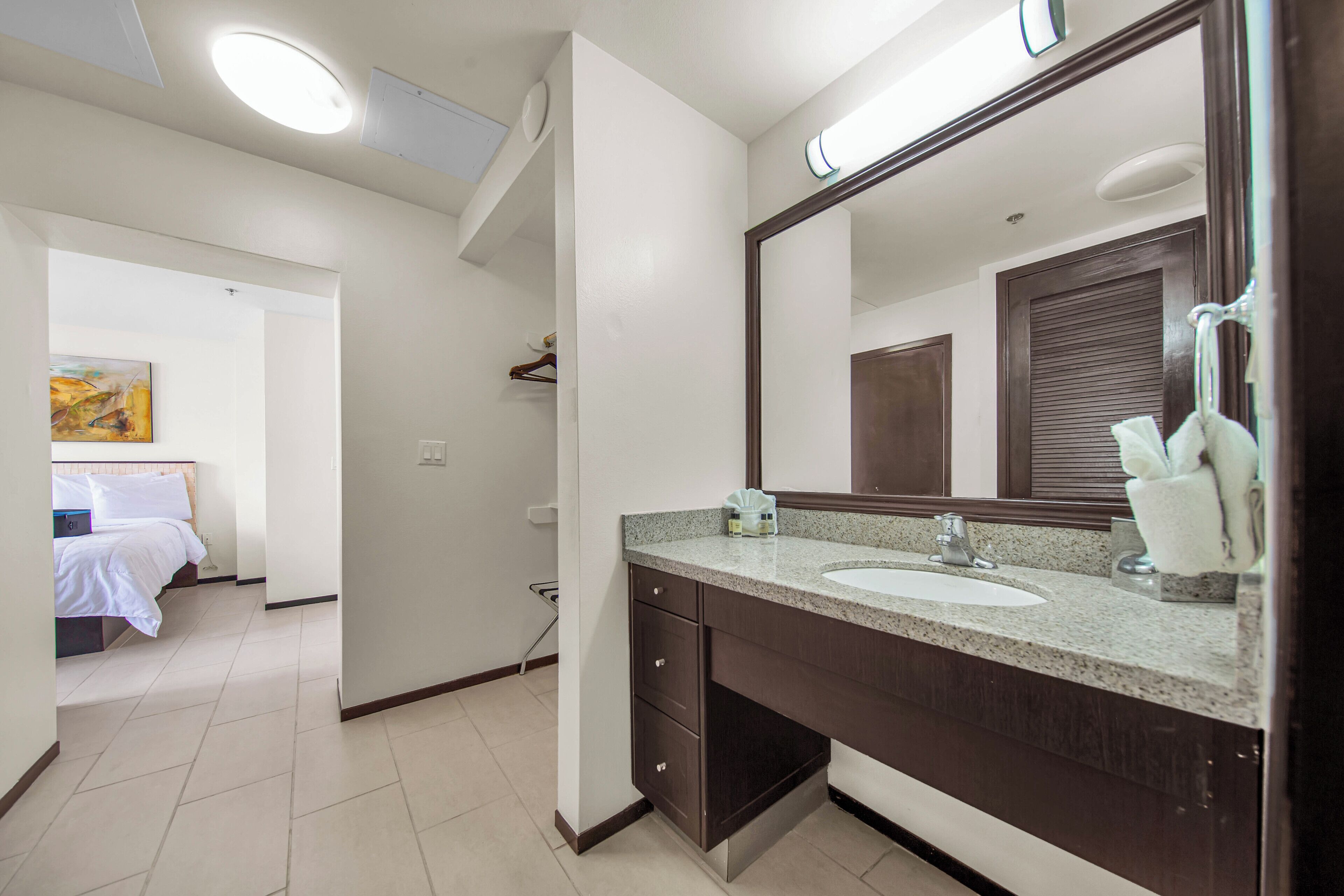 Une salle de bain d’hôtel avec un grand miroir, un lavabo et des toilettes. On peut apercevoir un lit dans la pièce adjacente.
