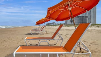 Een strand met rijen ligbedden en parasols, een heldere lucht en gebouwen op de achtergrond.