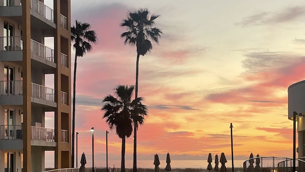 A swimming pool with a sunset view, palm trees, and a multi-story building with balconies.