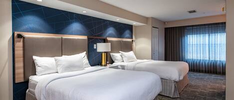 Habitación de hotel con dos camas, un ventanal grande con cortinas, un cabecero moderno y una alfombra estampada.