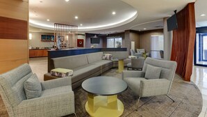 Un moderno vestíbulo de hotel con techo curvo, un área de estar central con sofás y sillones, una pequeña mesa redonda y una cocina al fondo.